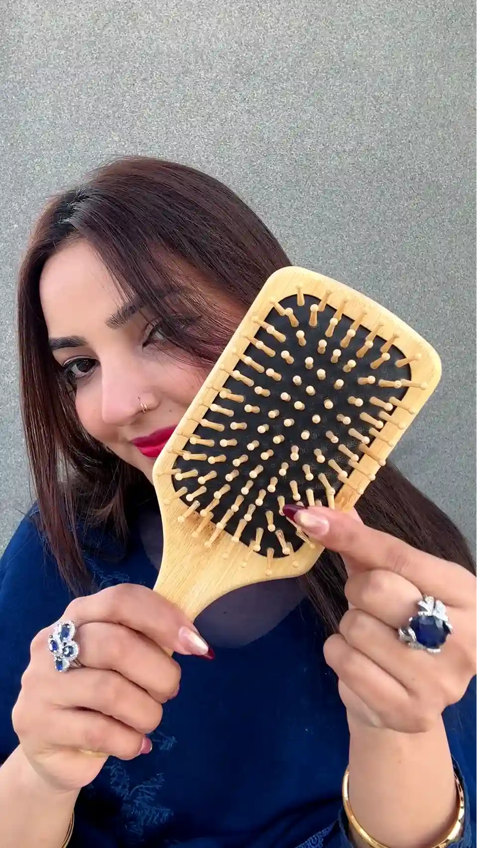 Just Amna wooden hairbrush held by hands with blue-stone rings, gray background
